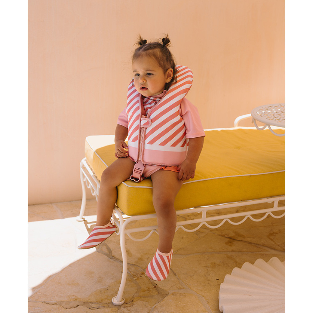 Criança vestindo colete salva-vidas rosa com riscas brancas sentada em banco com almofada amarela
