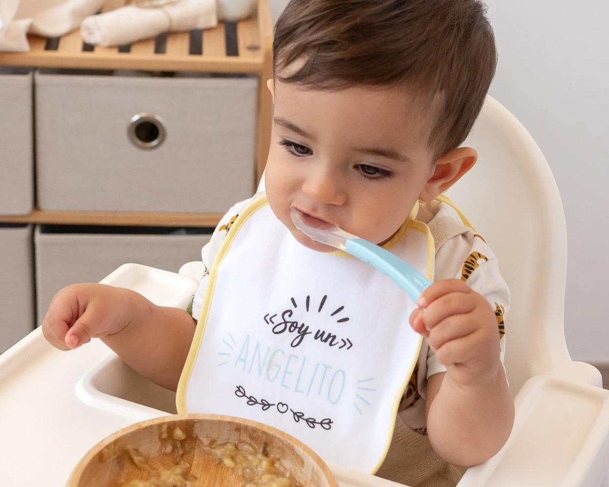 Bebé com babete sentado numa cadeira alta, com colher e tigela de papa.