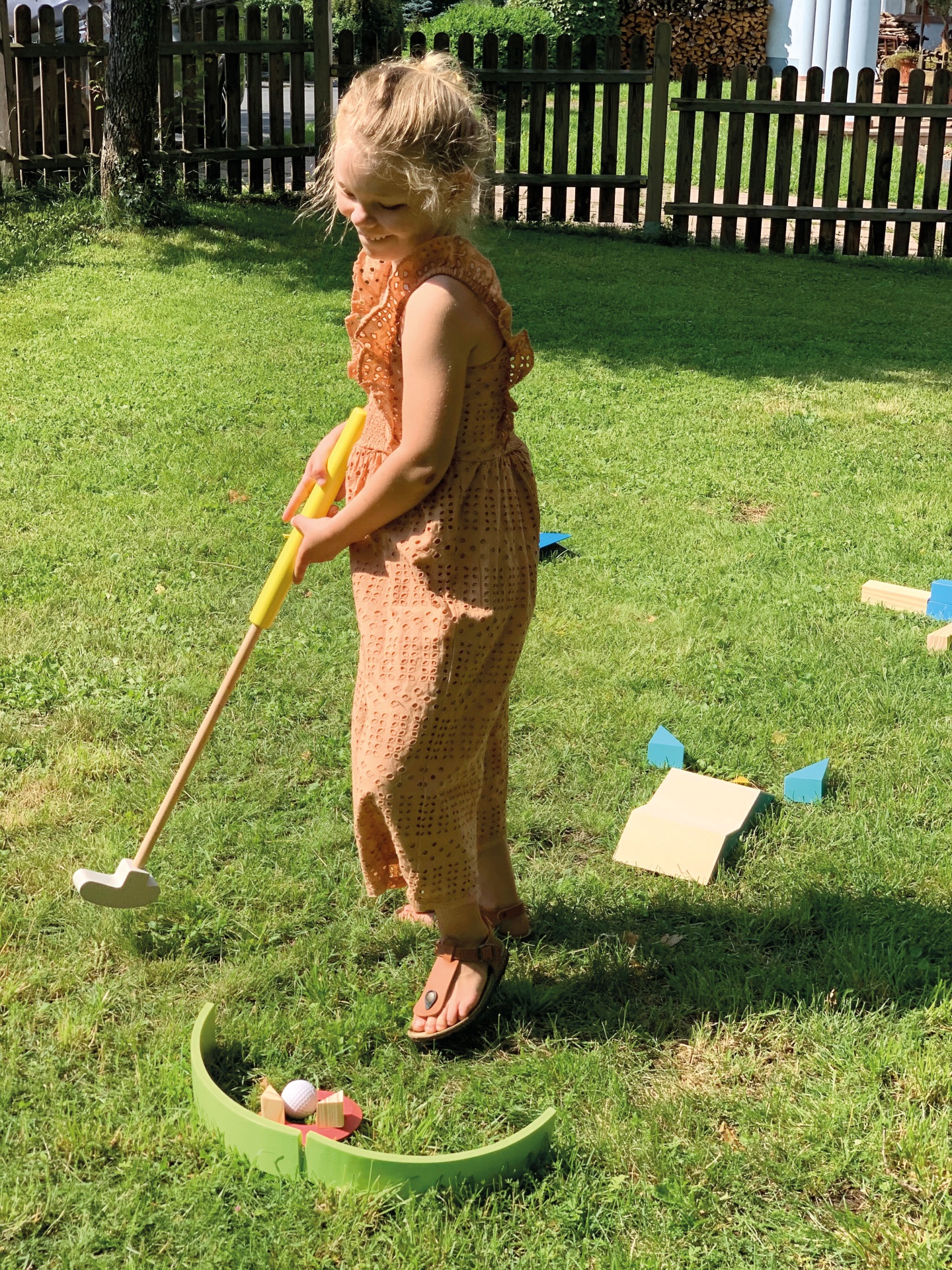 Menina a jogar mini-golfe num relvado com taco amarelo e vestido castanho