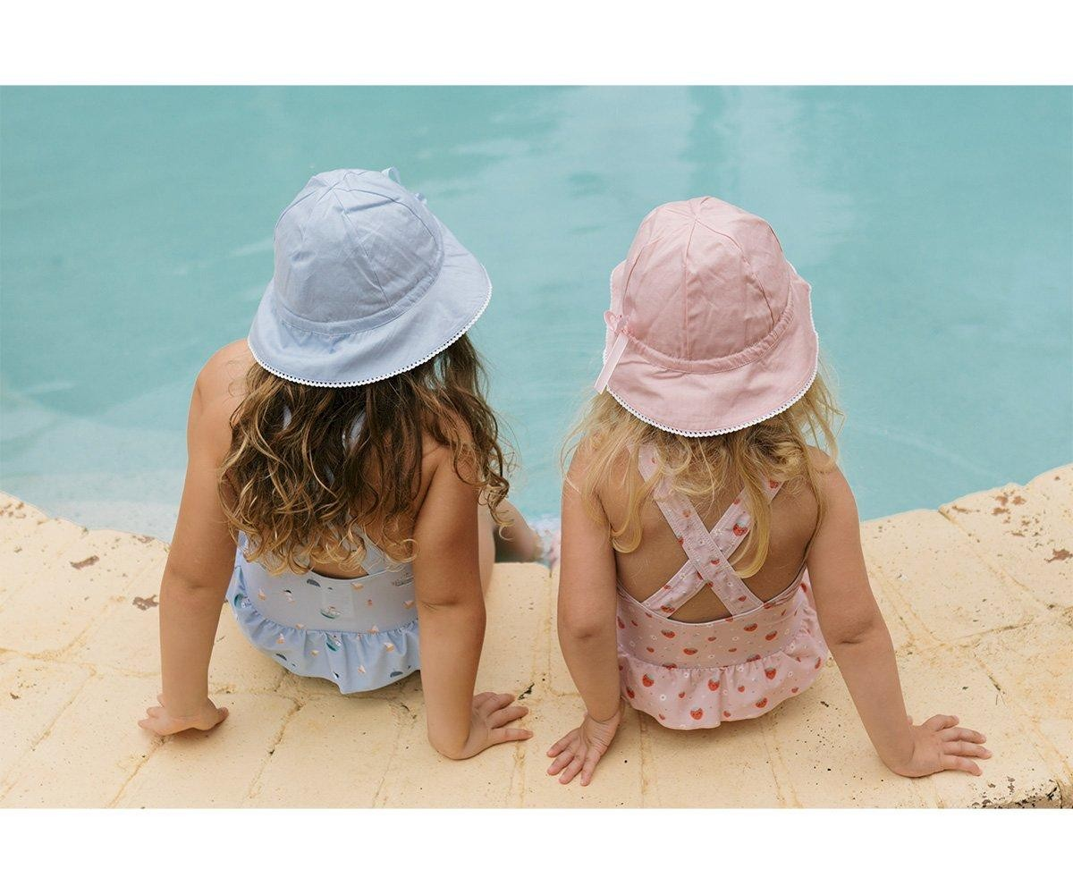 Duas crianças sentadas de costas junto a piscina, com fatos de banho e chapéus de balde azul e rosa.