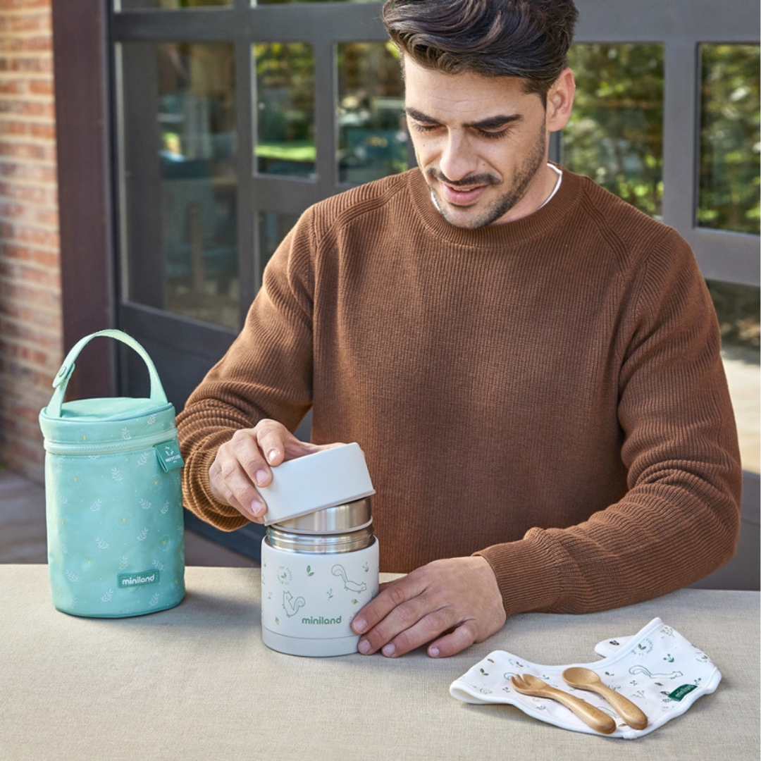 Homem a abrir lancheira térmica branca e verde com desenhos de esquilos, saco térmico e talheres de madeira sobre mesa.