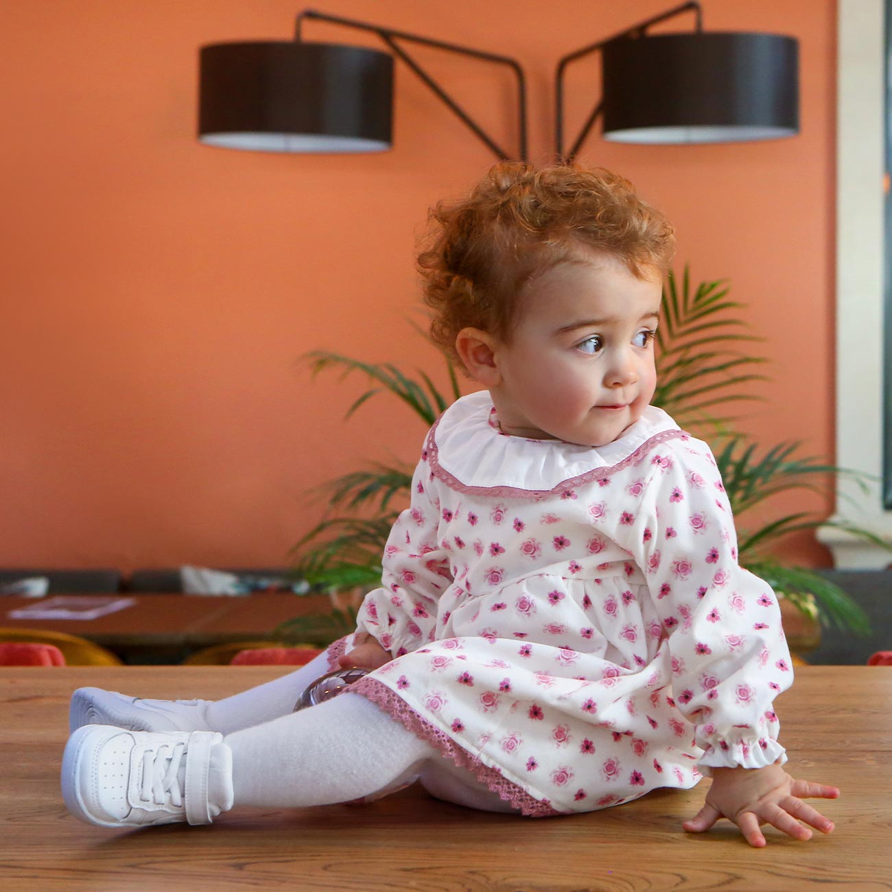 Bebé com vestido branco estampado sentada numa mesa de madeira com candeeiro preto e planta ao fundo.