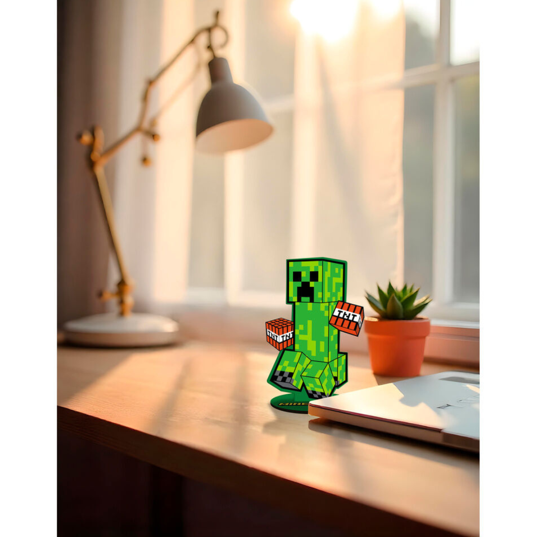 Figura decorativa de Creeper do Minecraft em cartão verde com caixas de TNT, sobre mesa de madeira junto a planta e portátil.