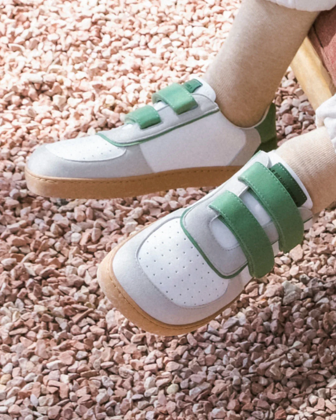 Sapatilhas brancas com detalhes verdes e sola castanha sobre pedras pequenas cor de rosa.