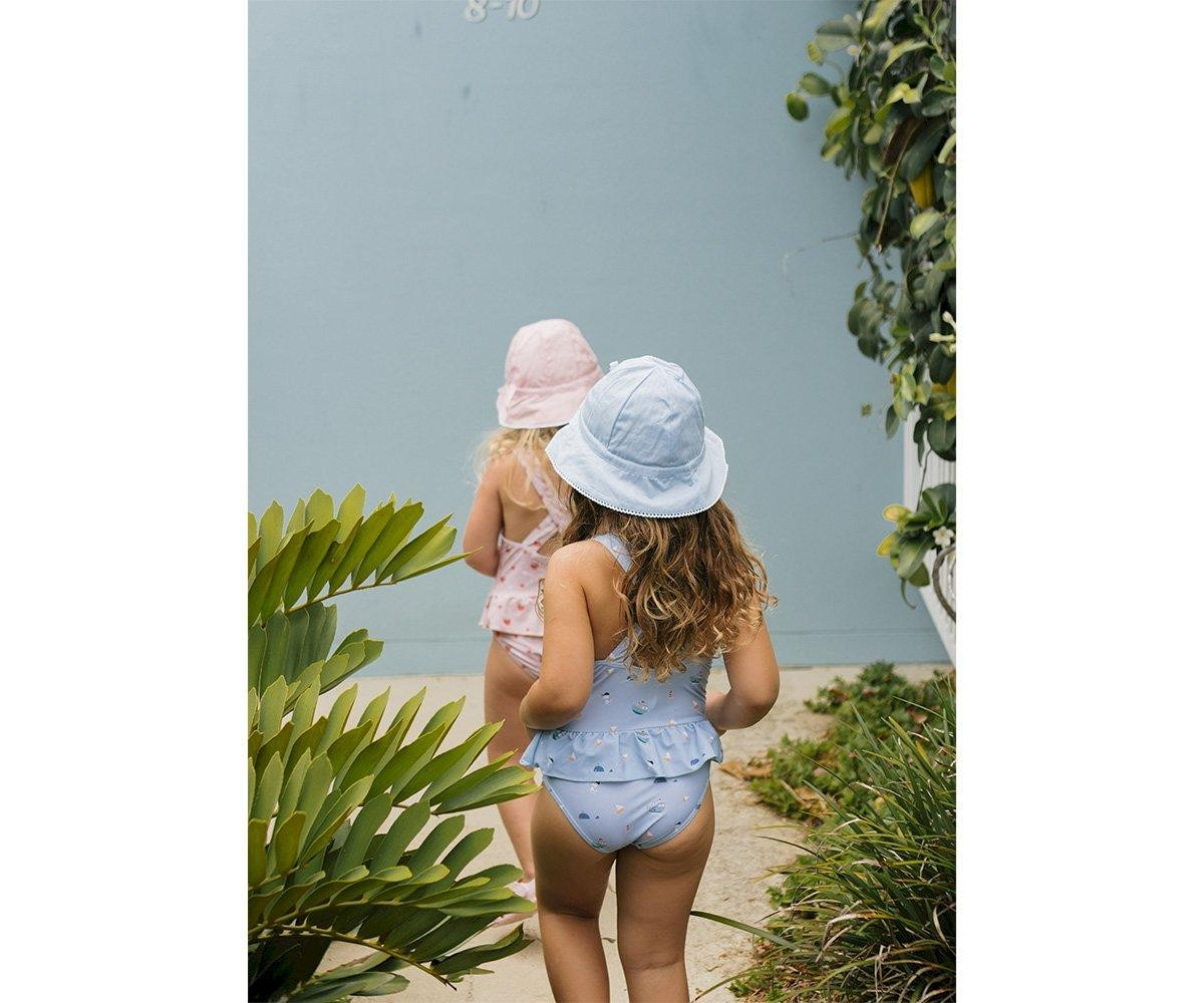 dois bebés com fatos de banho e chapéus, parede azul e plantas