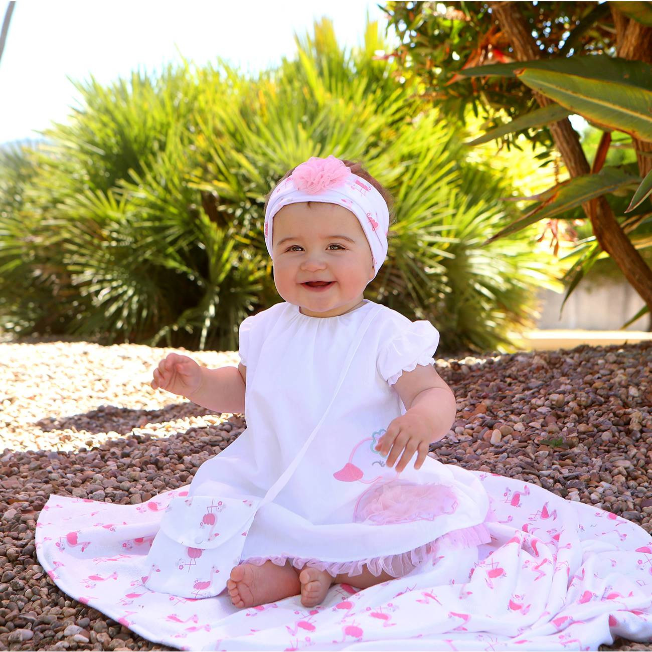 Bebé com vestido branco e rosa, sentada no chão de pedra com plantas verdes ao fundo