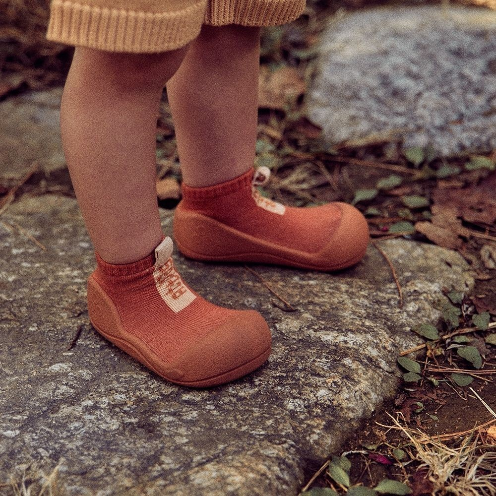 Sapatos para crianças terracota com etiqueta 'bobux' numa superfície de pedra