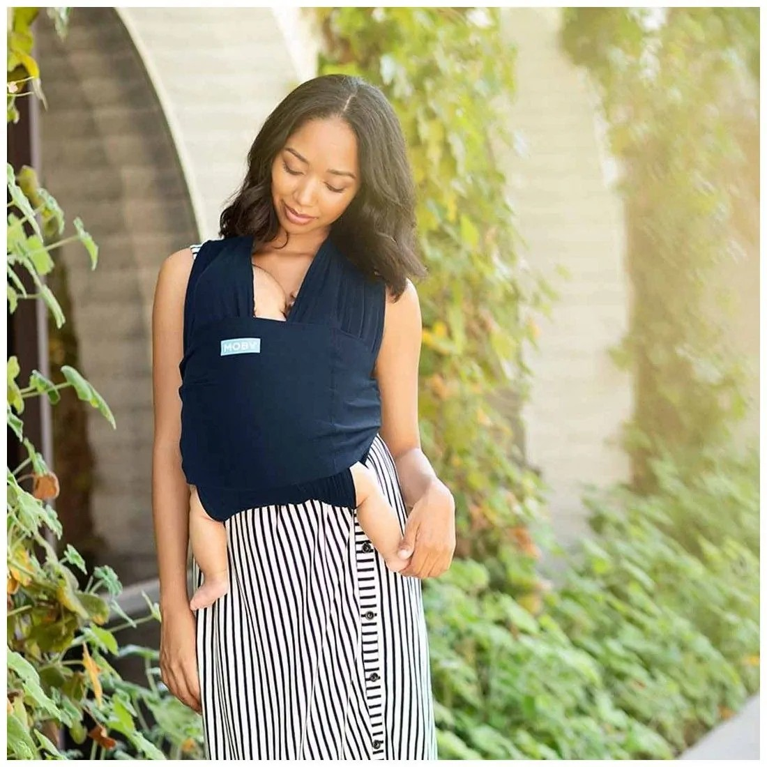 Mãe a usar canguru azul escuro com bebé em ambiente exterior com vegetação