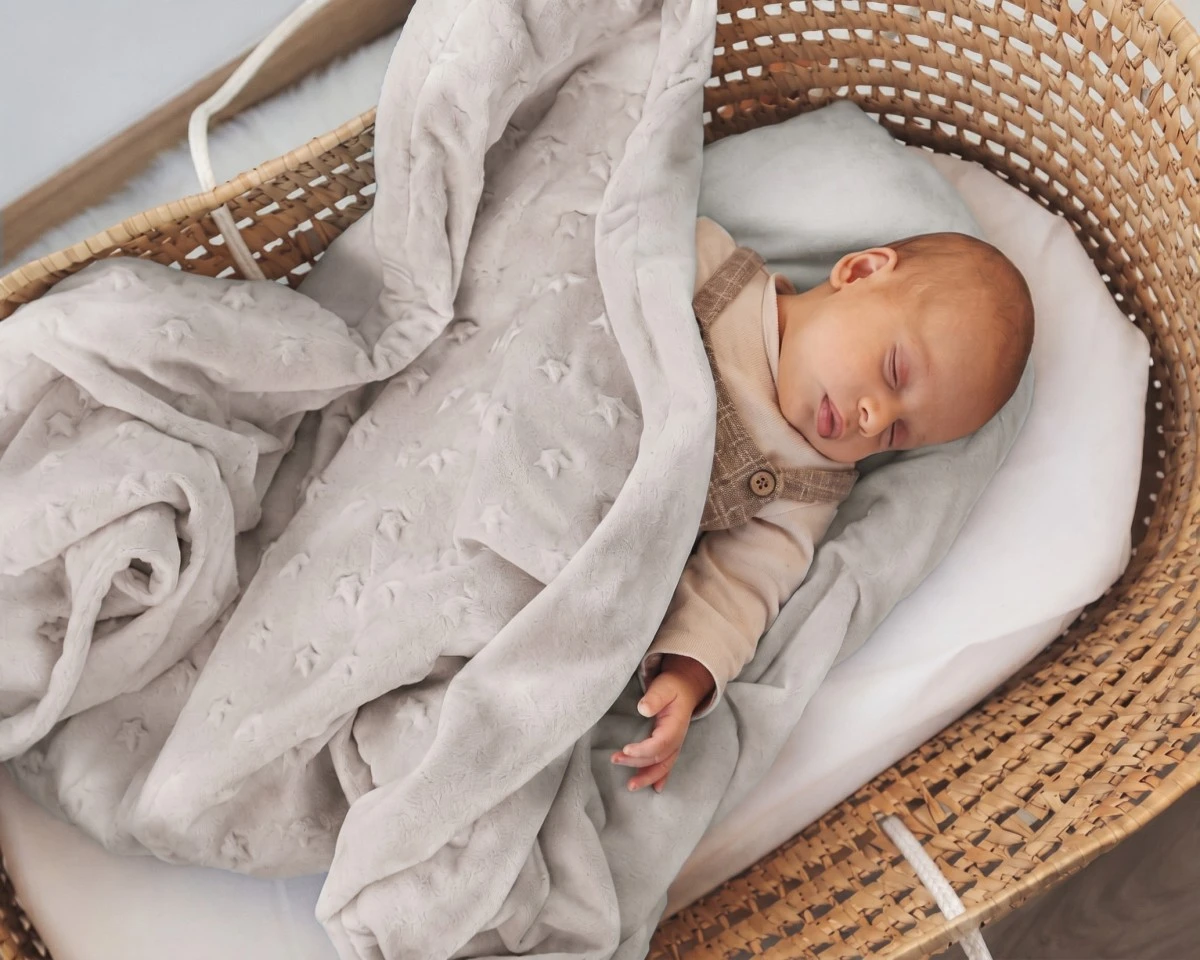 Bebé a dormir numa cama oval de verga com cobertor cinzento claro de estrelas em relevo