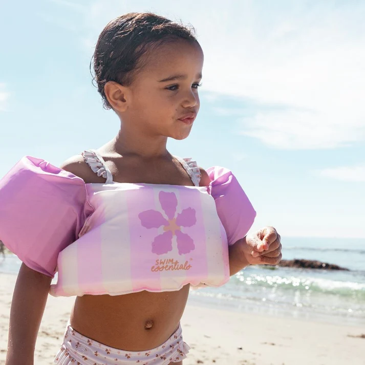 Criança com colete de natação cor de rosa e branco com flor, na praia com mar ao fundo