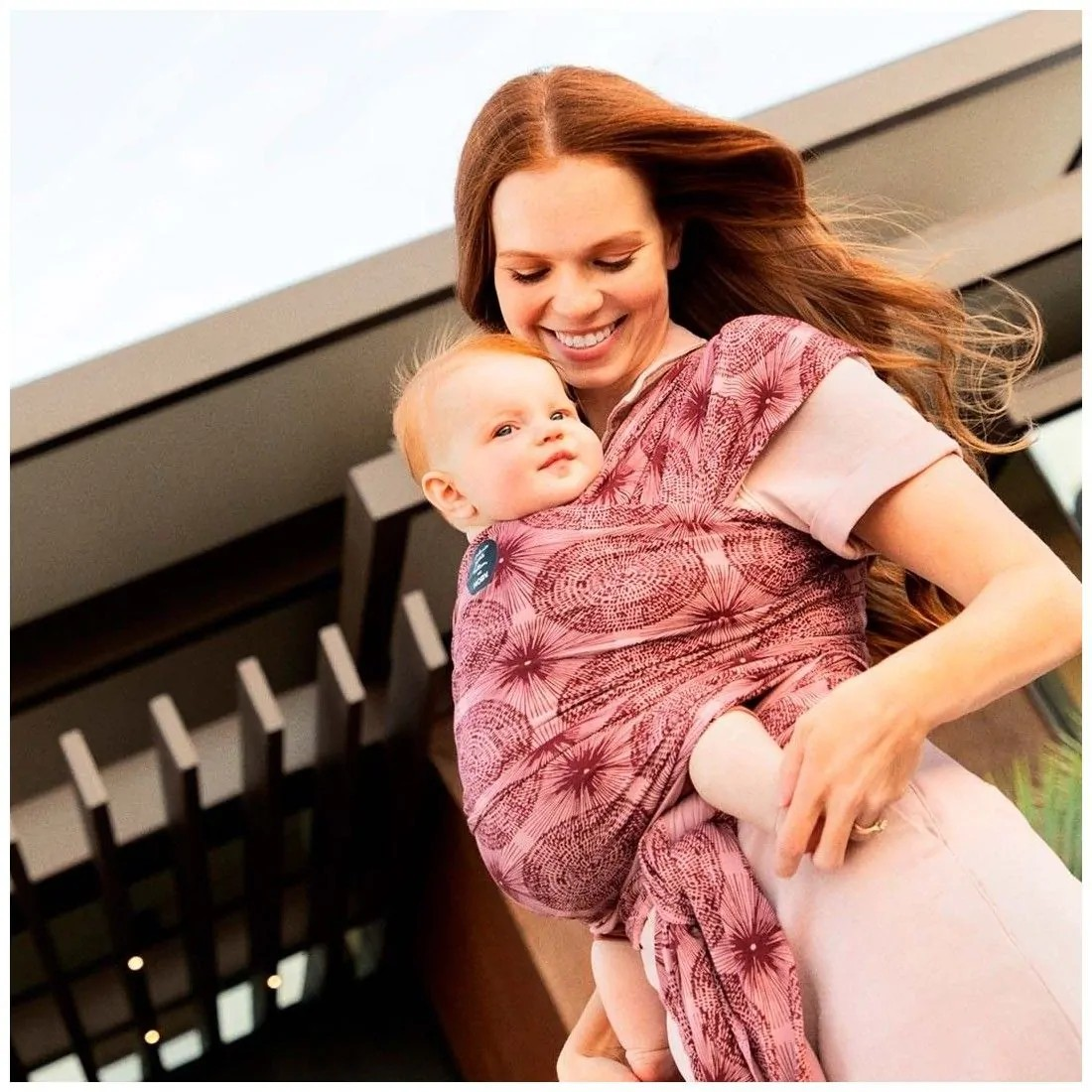 Mulher a usar porta-bebé em tecido cor de vinho com padrão geométrico a segurar bebé