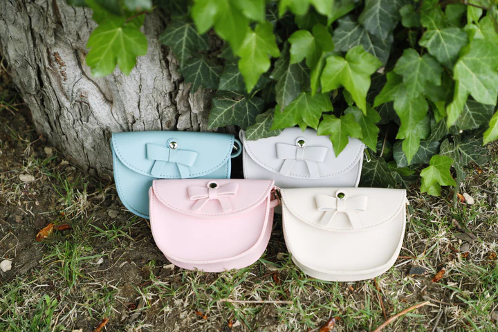 Quatro bolsas pequenas tipo carteira em cores pastel com laço decorativo perante um tronco de árvore e relva.