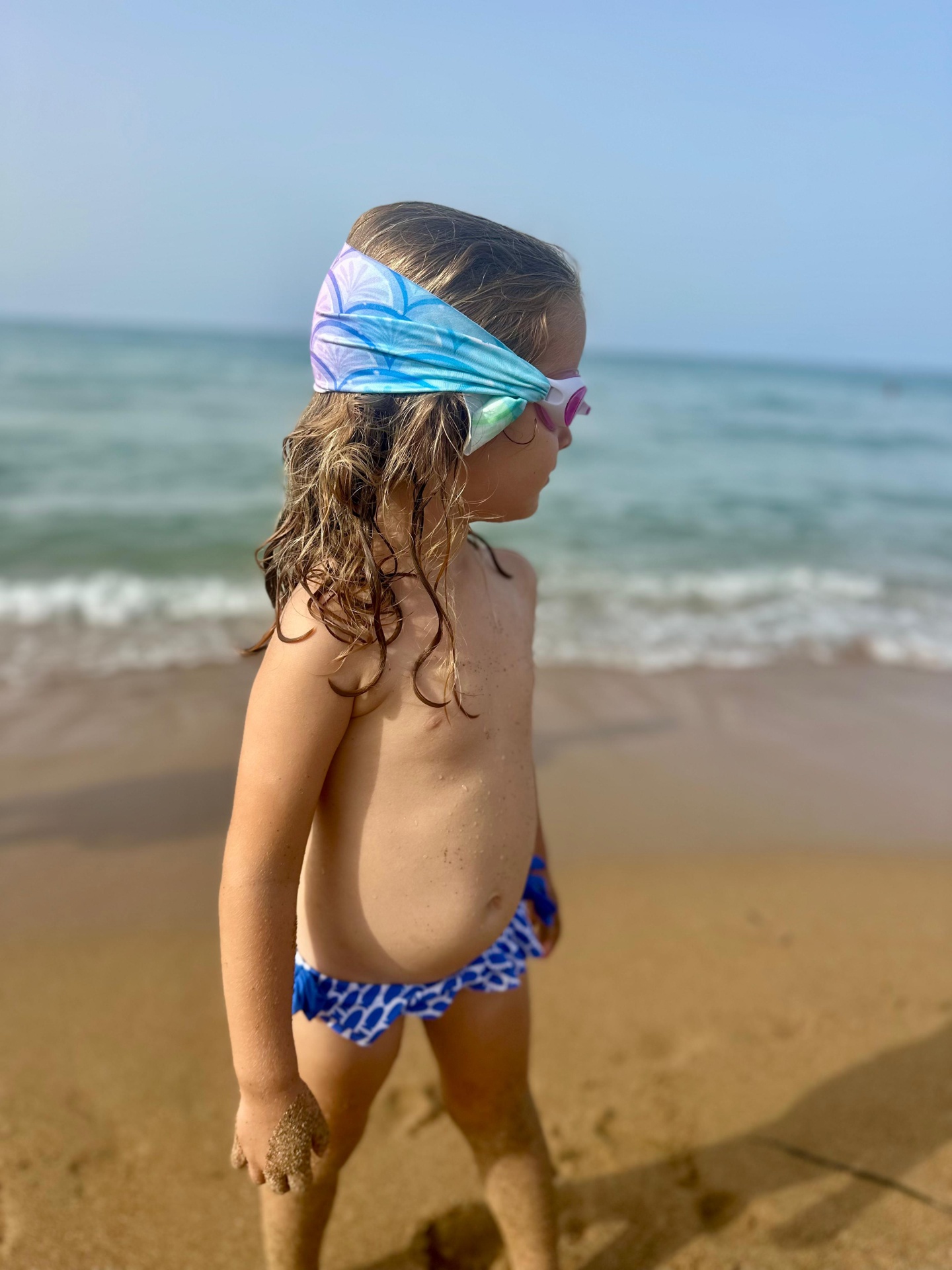Criança com óculos de natação cor-de-rosa e faixa de cabeça azul e roxa na praia