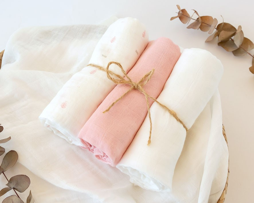 Três panos de tecidos enrolados, branco, rosa claro e branco com padrão, sobre pano branco maior com folhas secas.