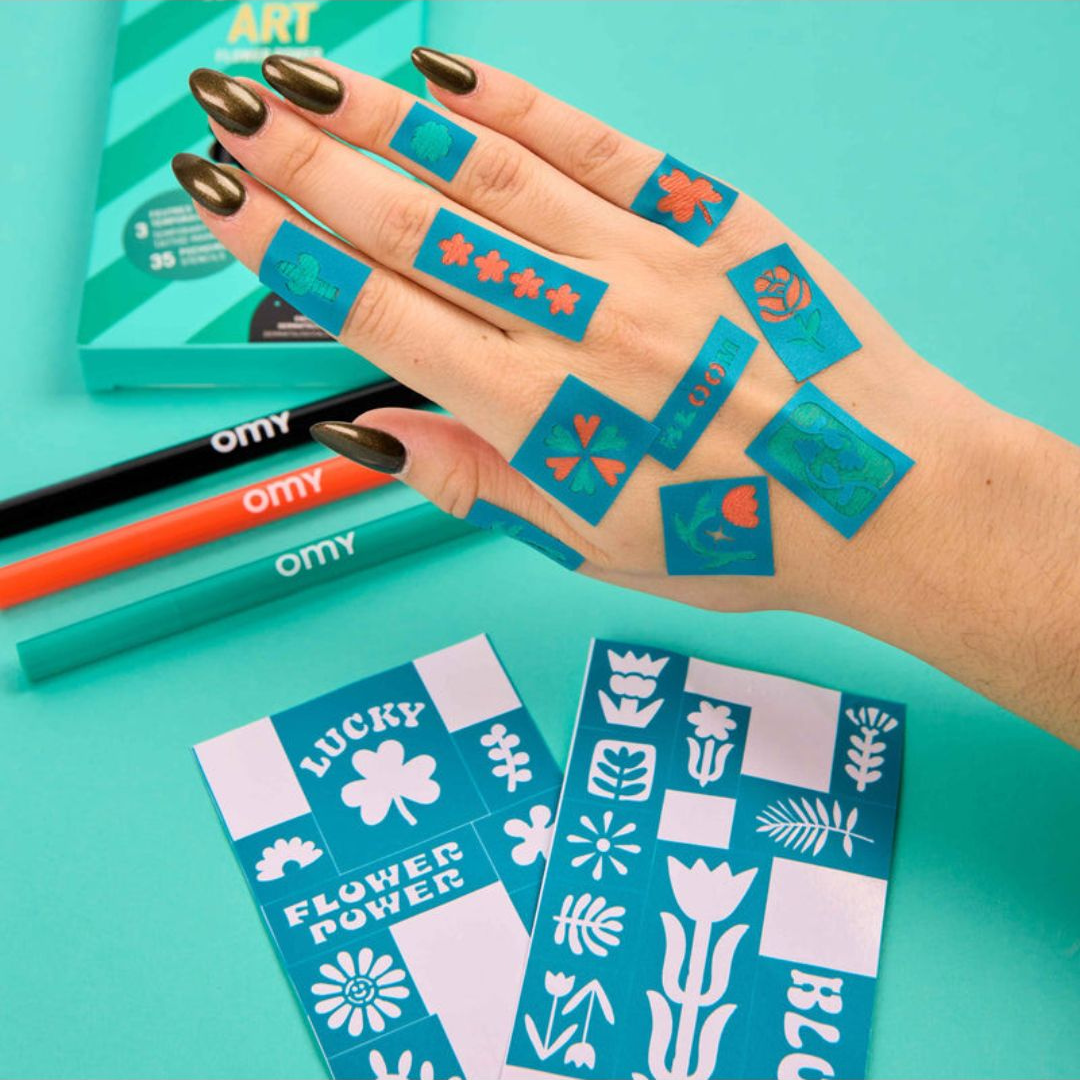 Mão com tatuagens temporárias azuis com desenhos florais e símbolos sobre fundo azul com lápis coloridos e folhas decorativas