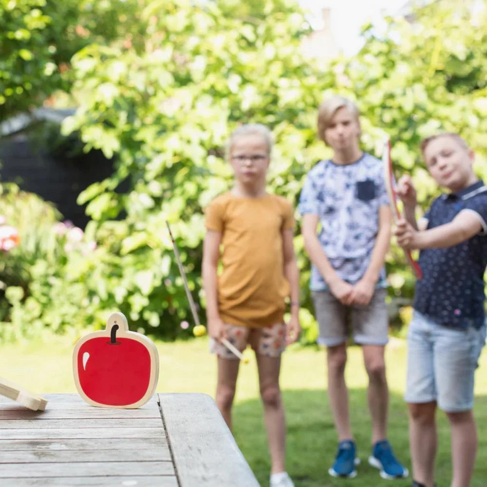 Jogo de dardos com alvo em forma de maçã num jardim com crianças ao fundo