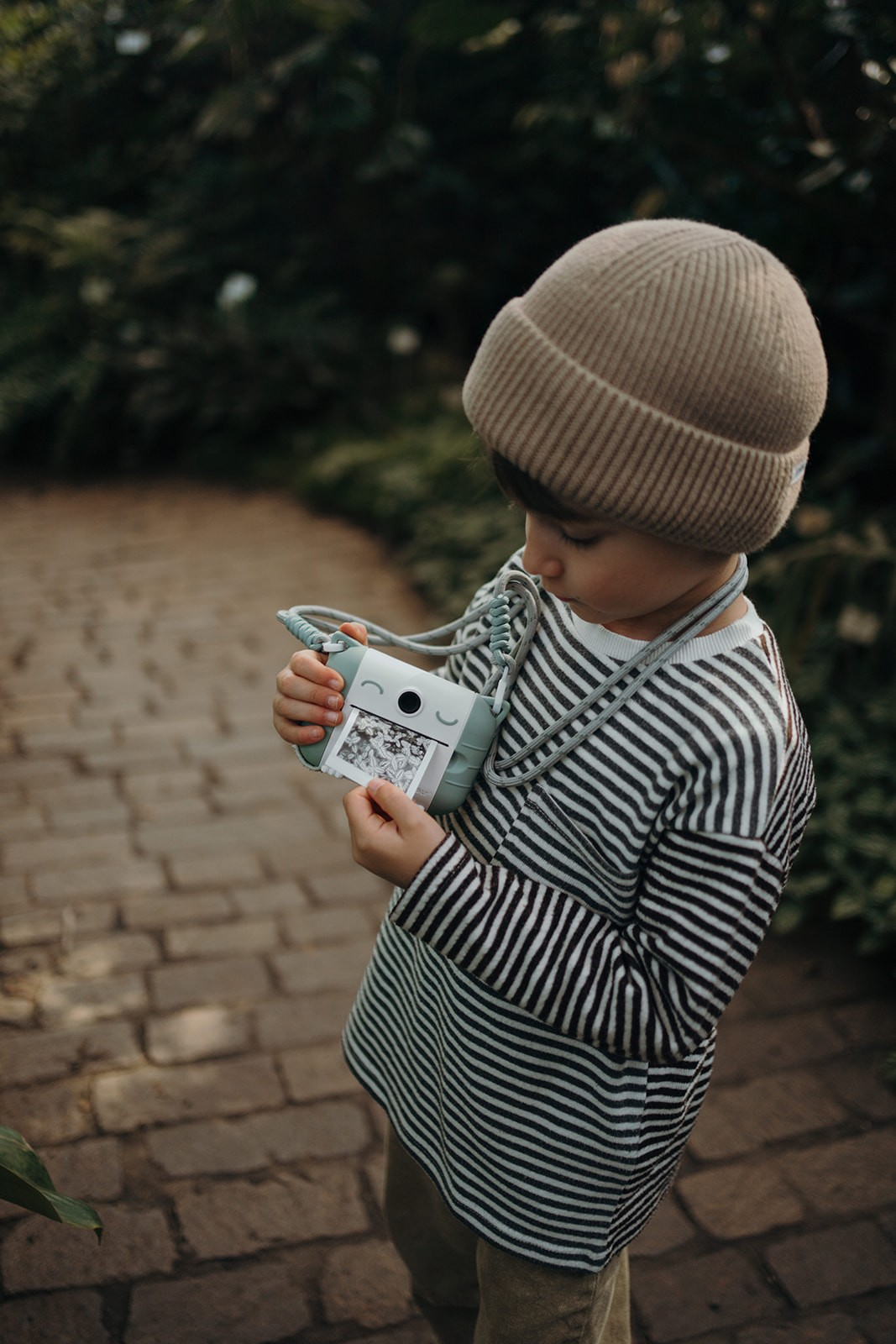 Criança com gorro e camisola de riscas segura câmara fotográfica cinza com rosto e tira