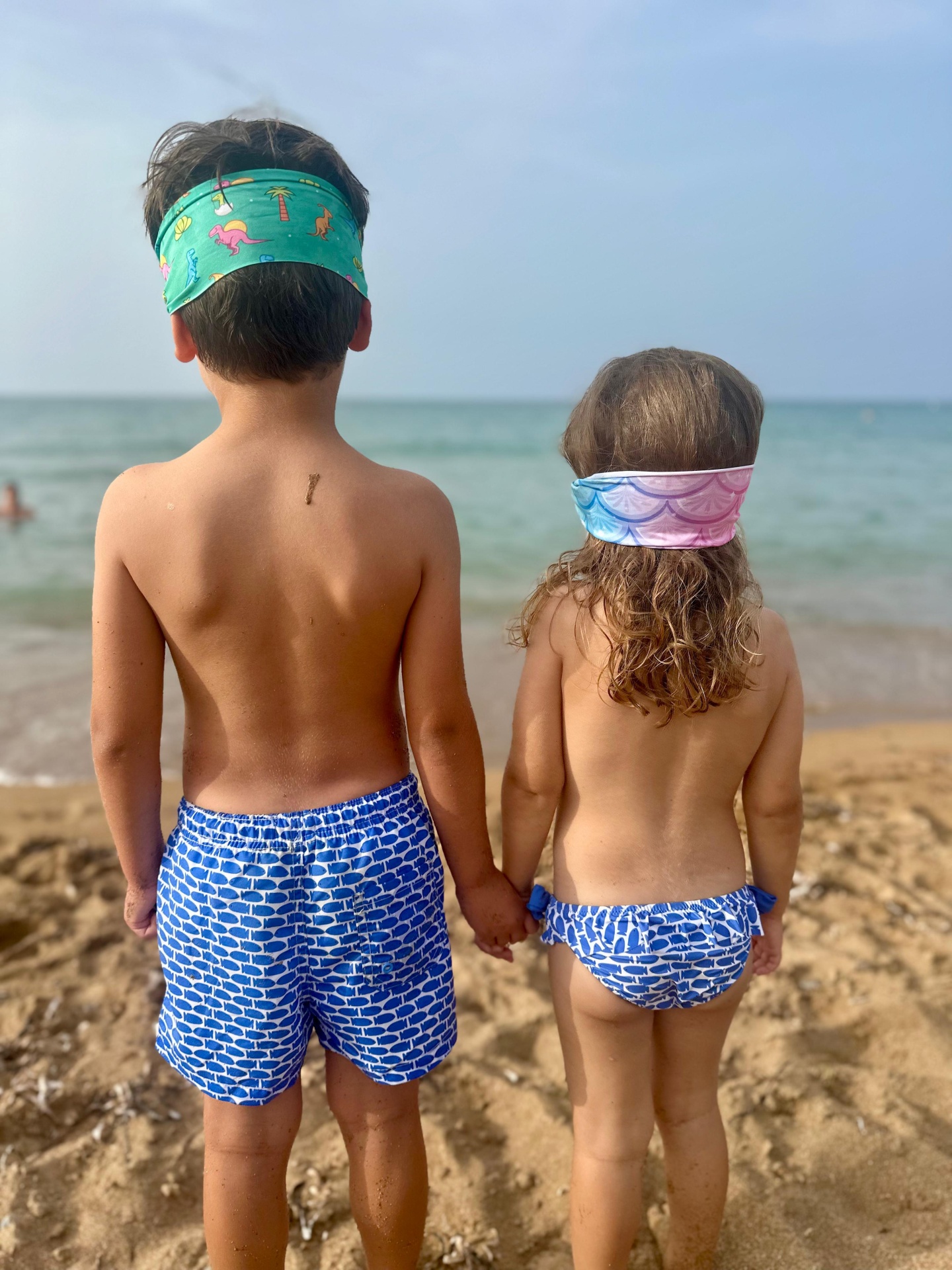 Crianças na praia com calções e biquíni azuis com padrão de peixes e lenços coloridos na cabeça.