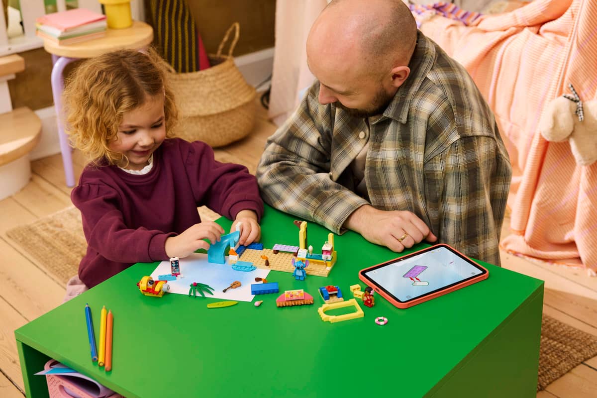 Mesa verde com peças de lego, lápis de cor e tablet com criança e adulto brincando