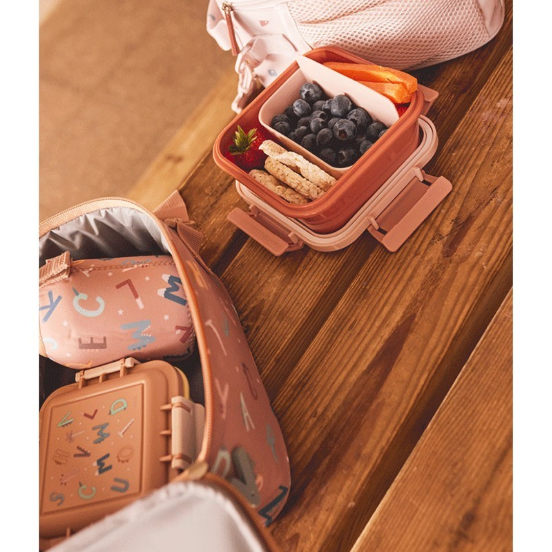 Recipientes de comida em tons de rosé e bege com frutas e bolachas numa mesa de madeira ao lado de uma bolsa rosada com letras coloridas