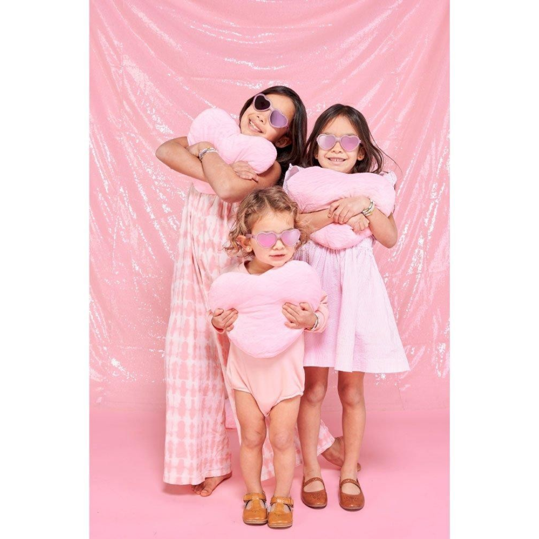 Três crianças com roupas e óculos de sol cor-de-rosa segurando almofadas macias rosa em fundo rosa brilhante