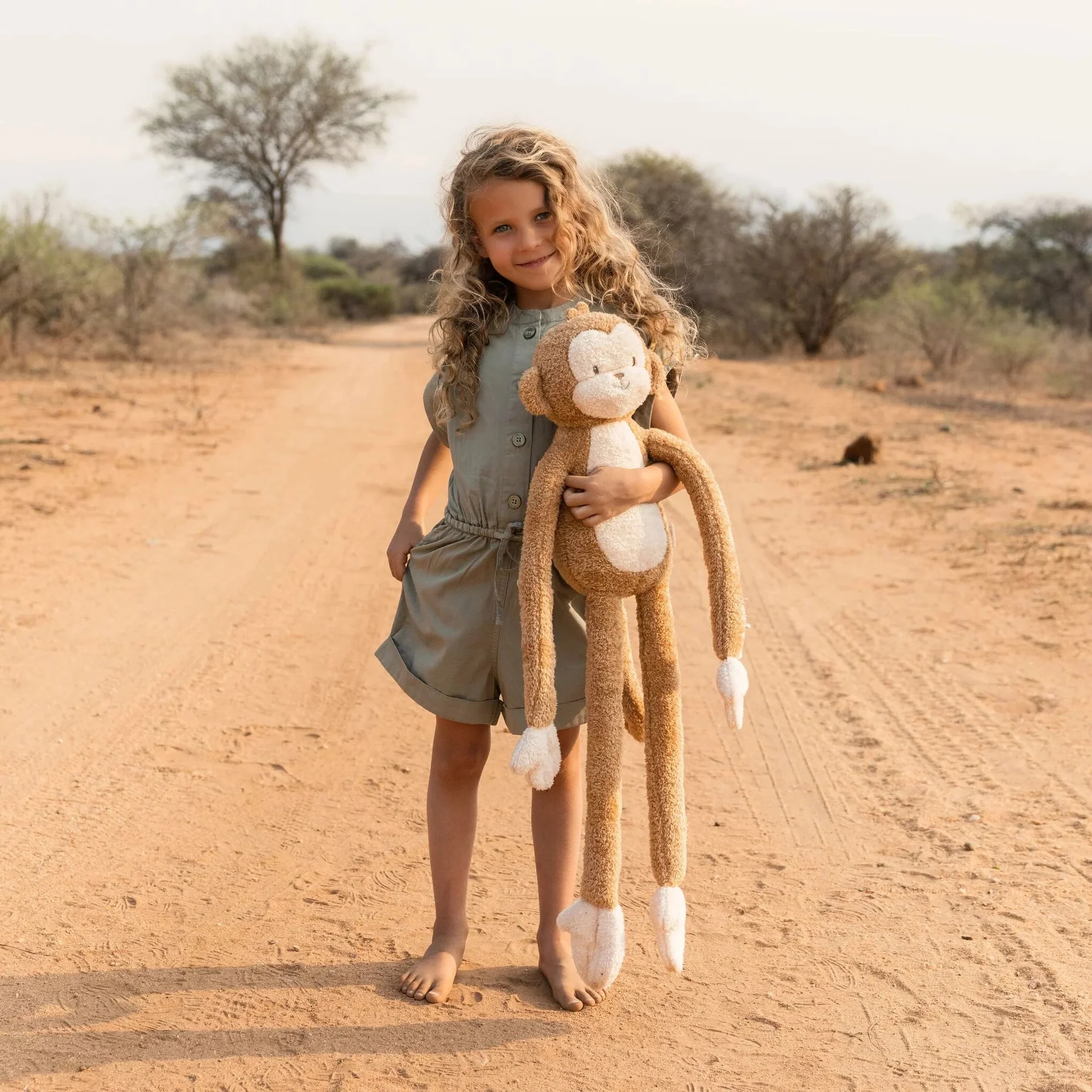 Menina com peluche de macaco numa estrada de terra batida com árvores secas ao fundo