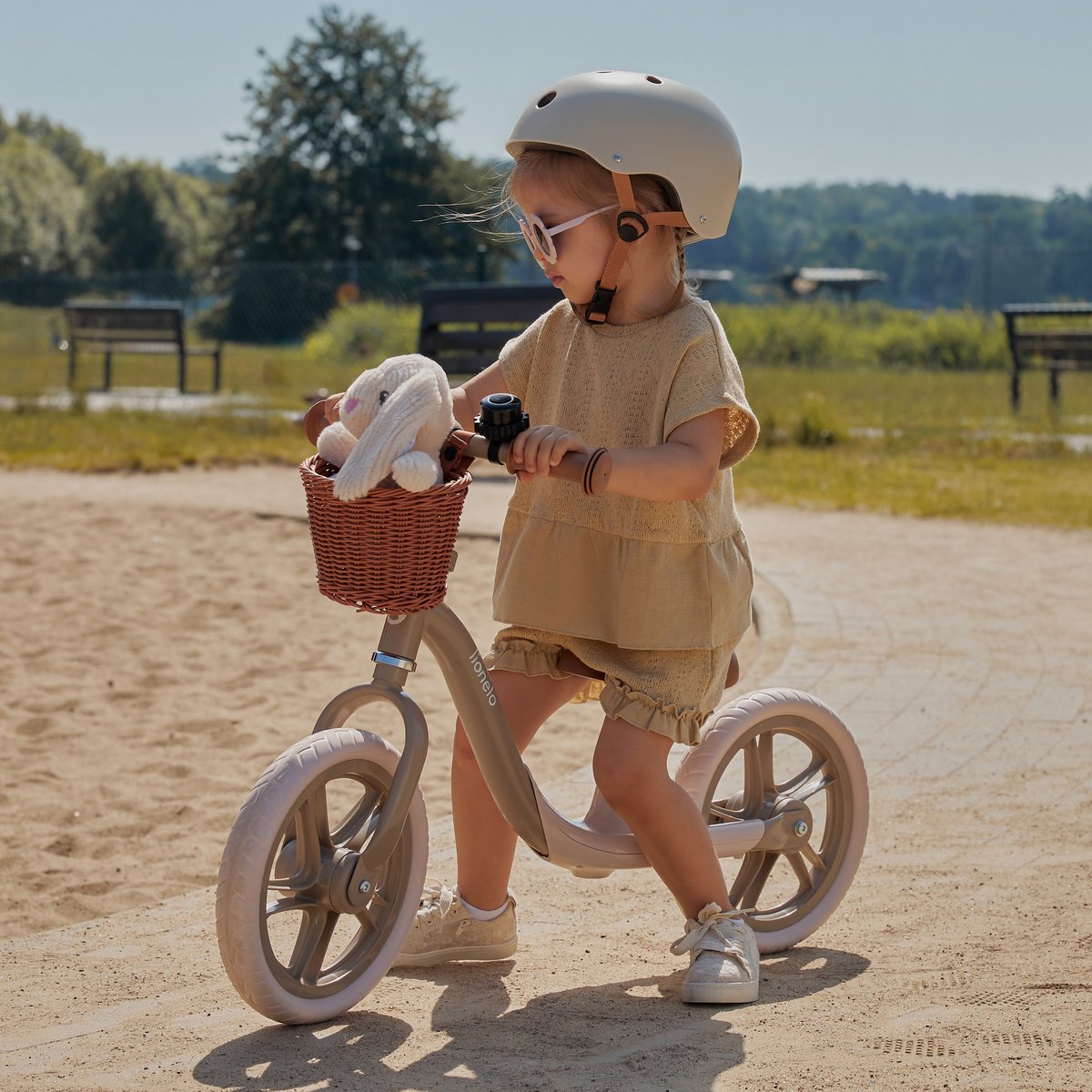 Criança montada numa bicicleta de equilíbrio cinza com capacete branco e brinquedo de peluche na cesta de vime