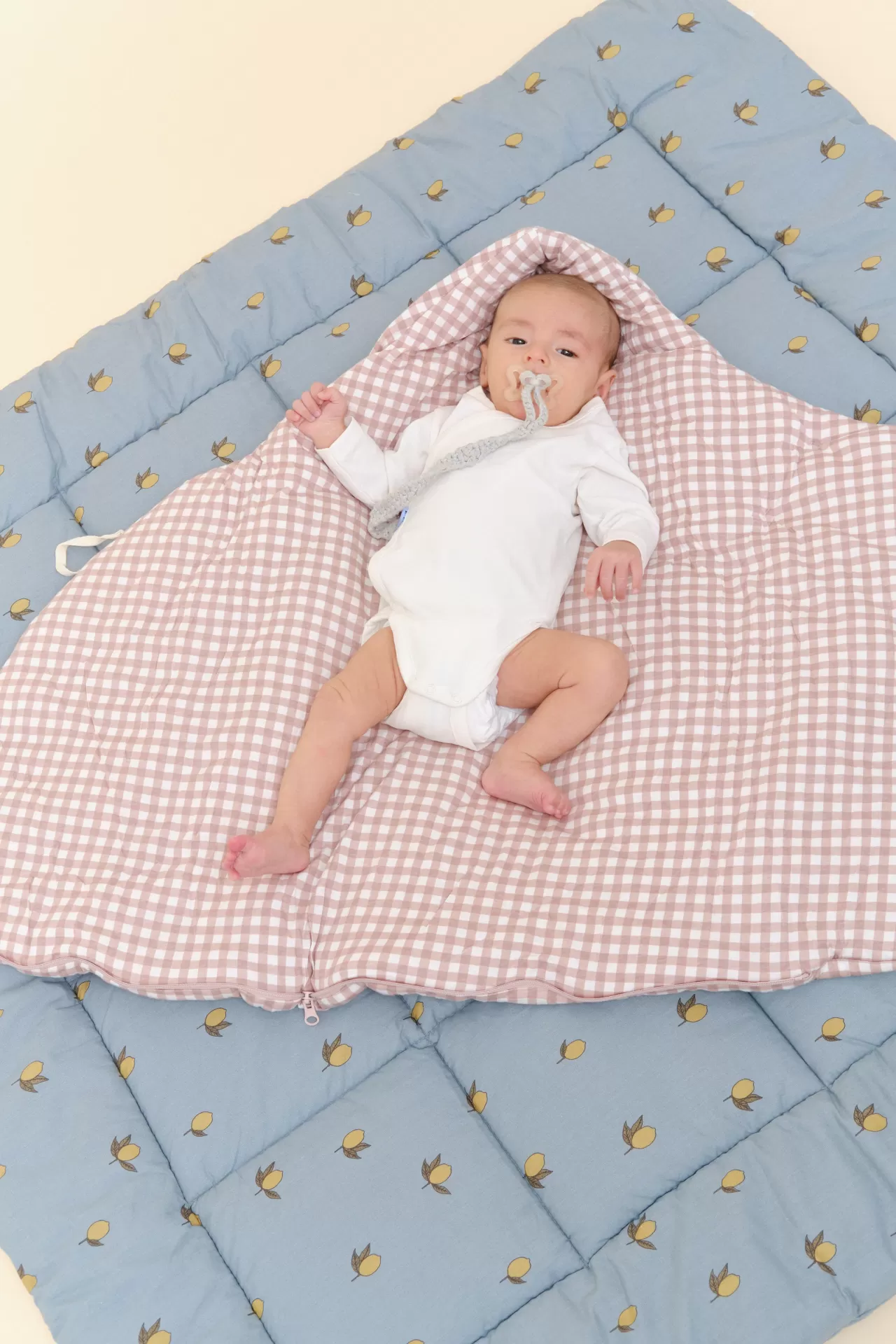 Bebé de body branco num saco de dormir xadrez rosa e branco sobre mantinha azul almofadada com limões.