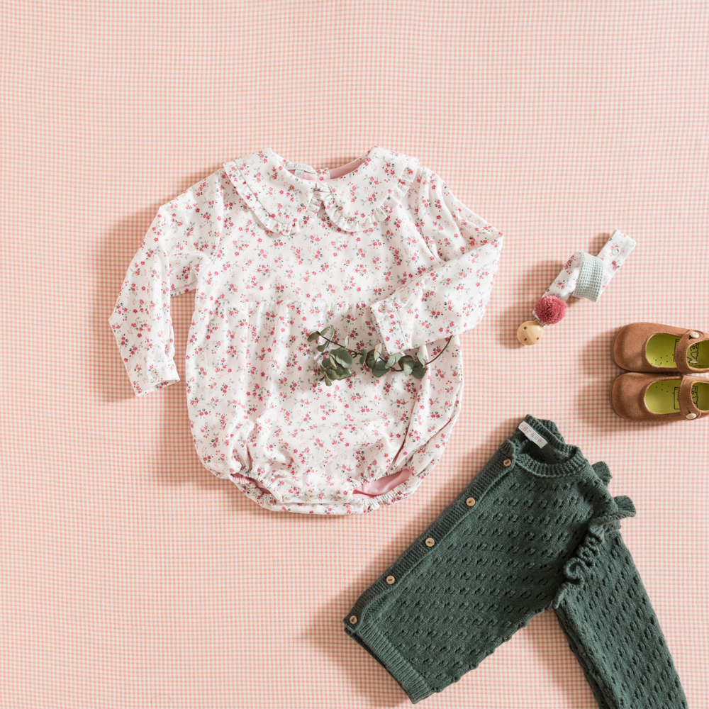 Conjunto de roupa de bebé com body floral branco, calças verdes de malha, sapatos castanhos e cinto colorido sobre fundo rosa xadrez