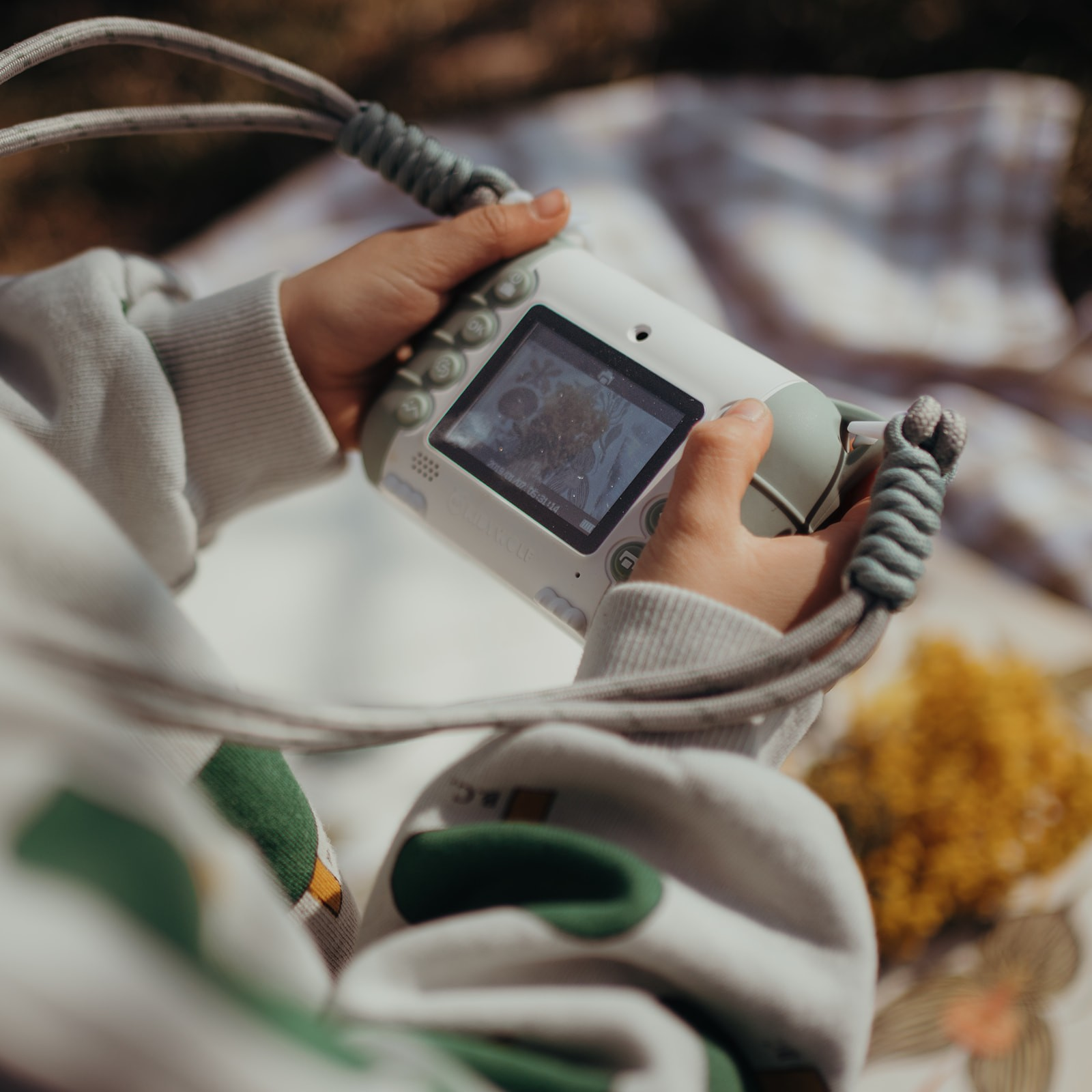 Câmara digital portátil branca com visor e pulseira cinzenta em mãos junto a tecido xadrez e flores