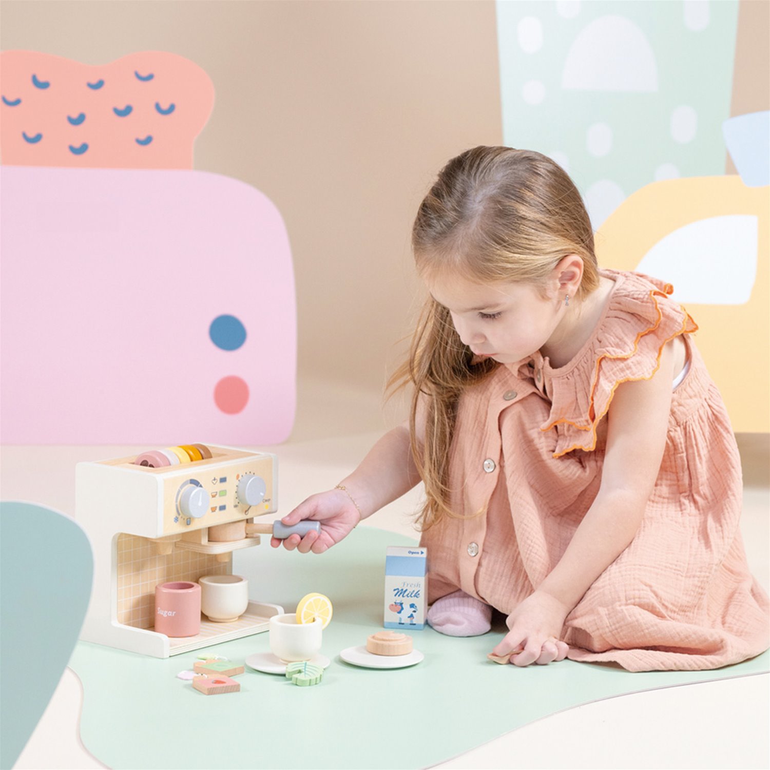 Menina com vestido salmão a brincar com máquina de café e acessórios de brinquedo feitos de madeira.