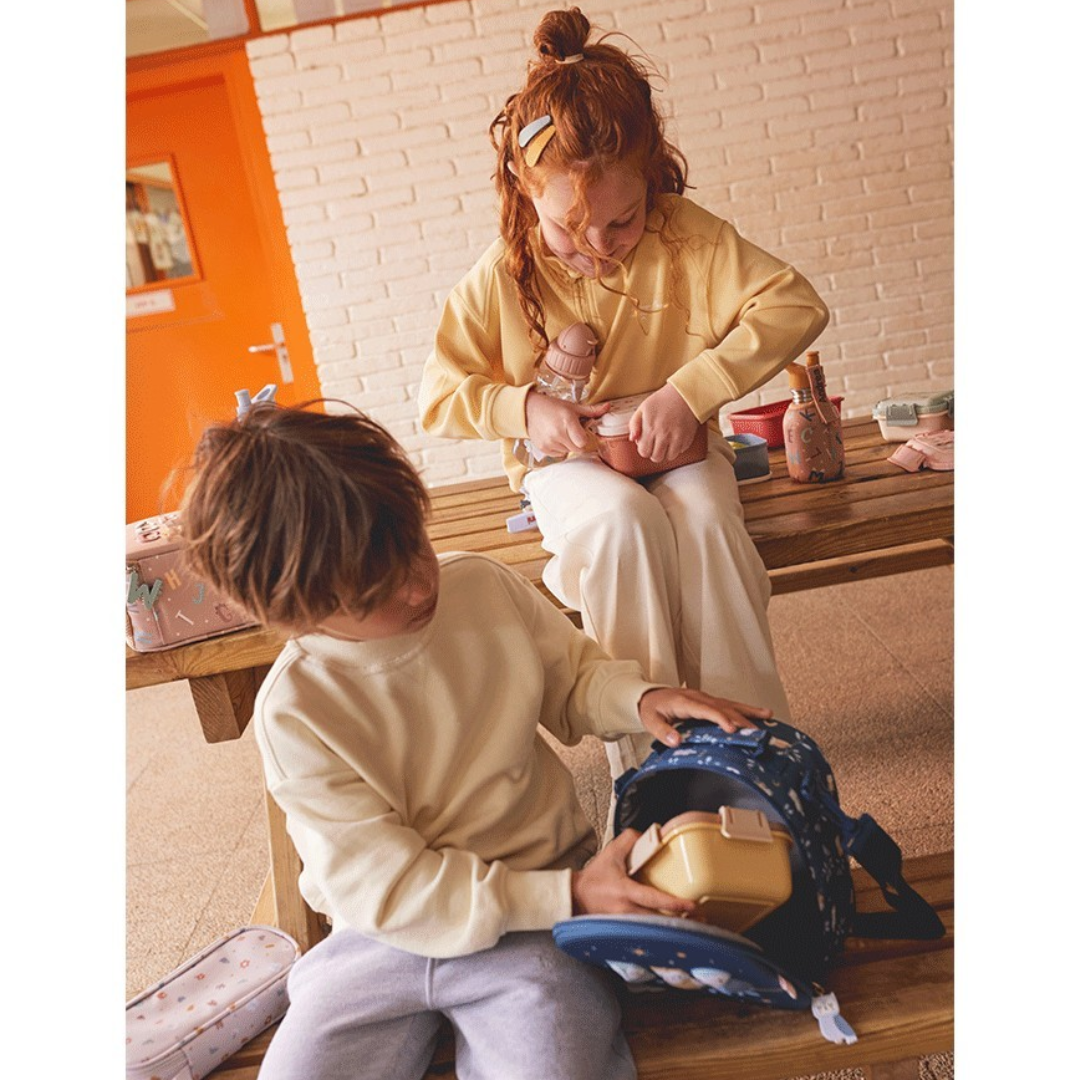 Criança com mochila azul e outra pessoa com camisola amarela clara sentadas numa mesa de madeira