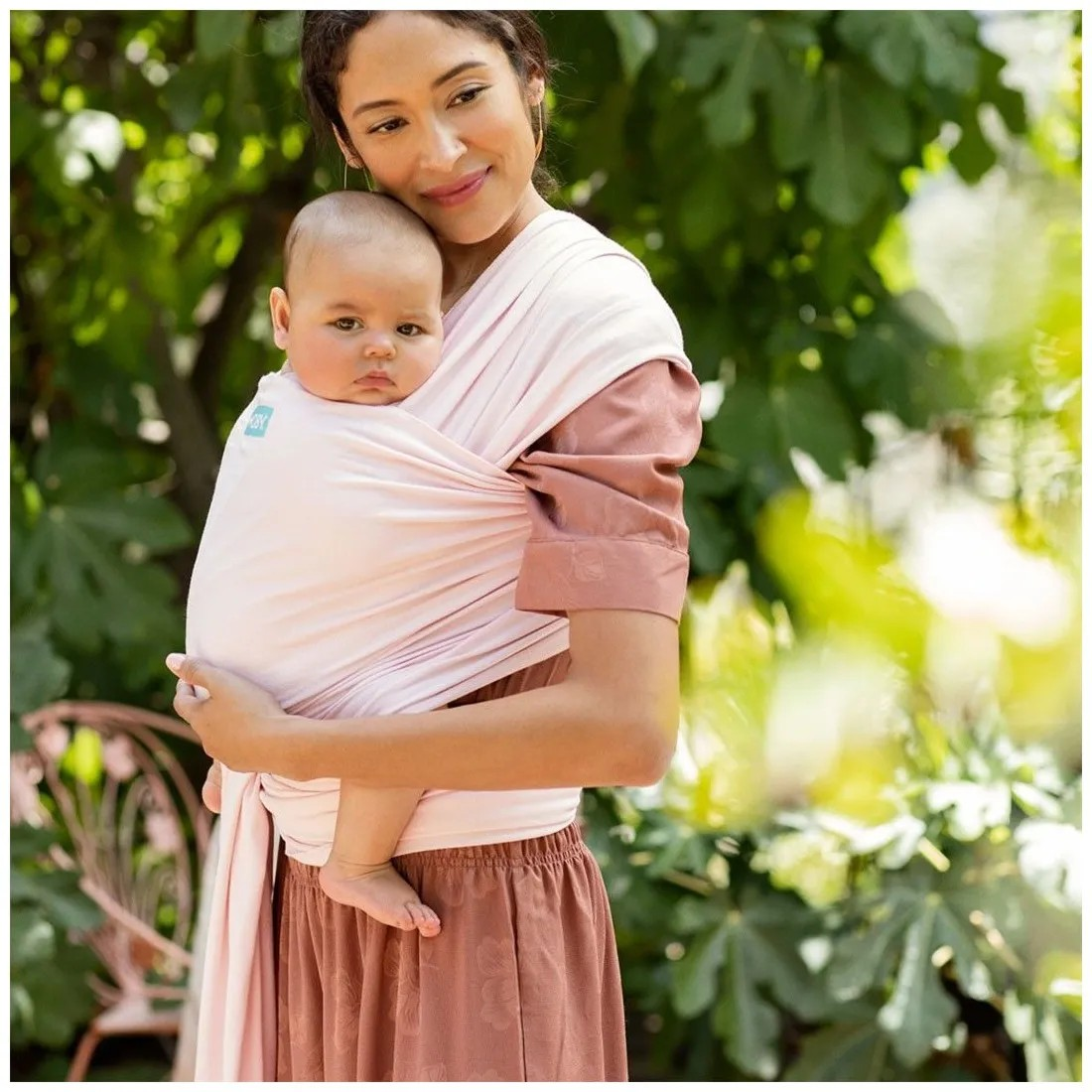 Mãe a usar sling cor de rosa claro com bebé ao colo, ao ar livre com vegetação
