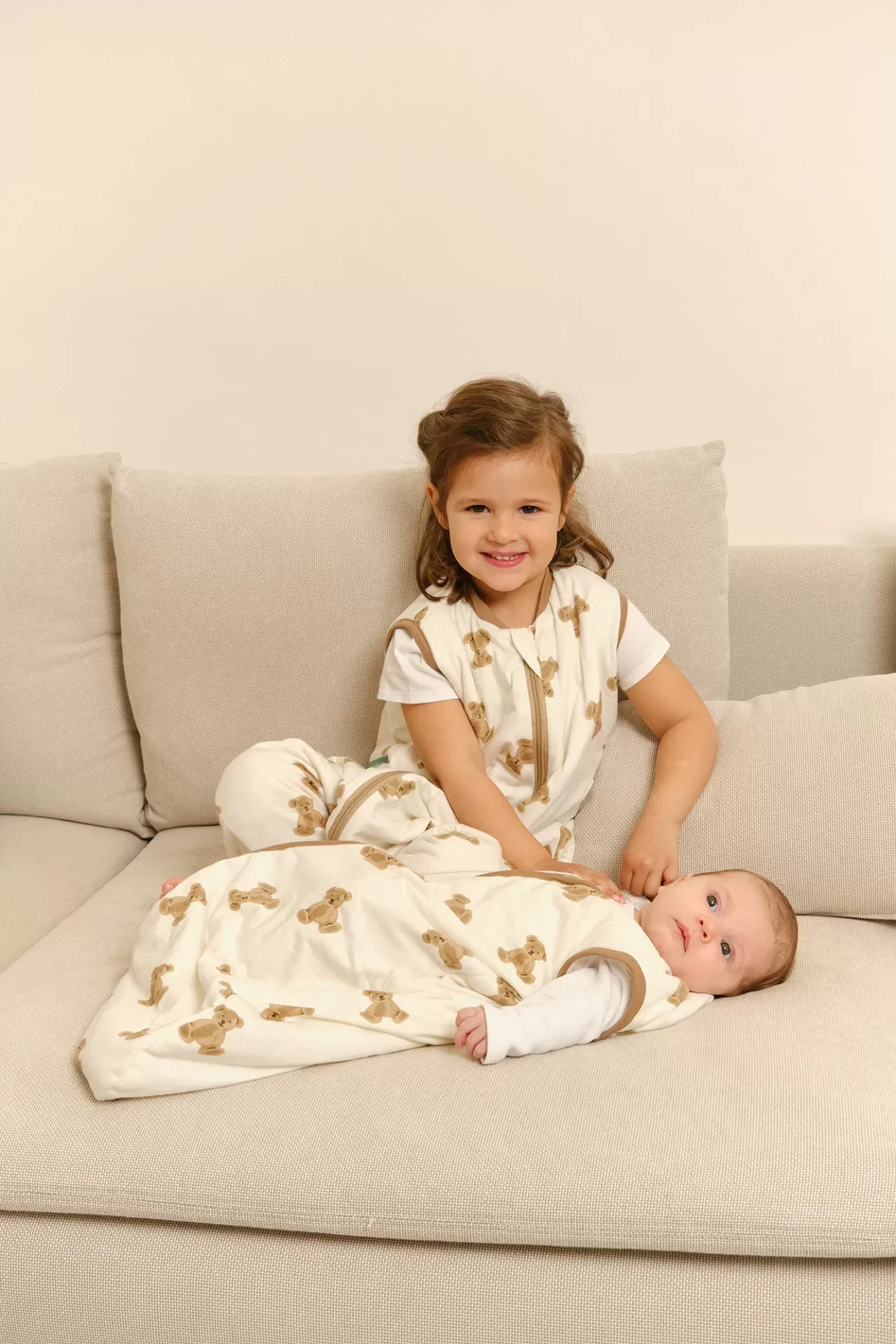 Duas crianças com sacos de dormir com padrão de ursinhos castanhos num sofá bege.