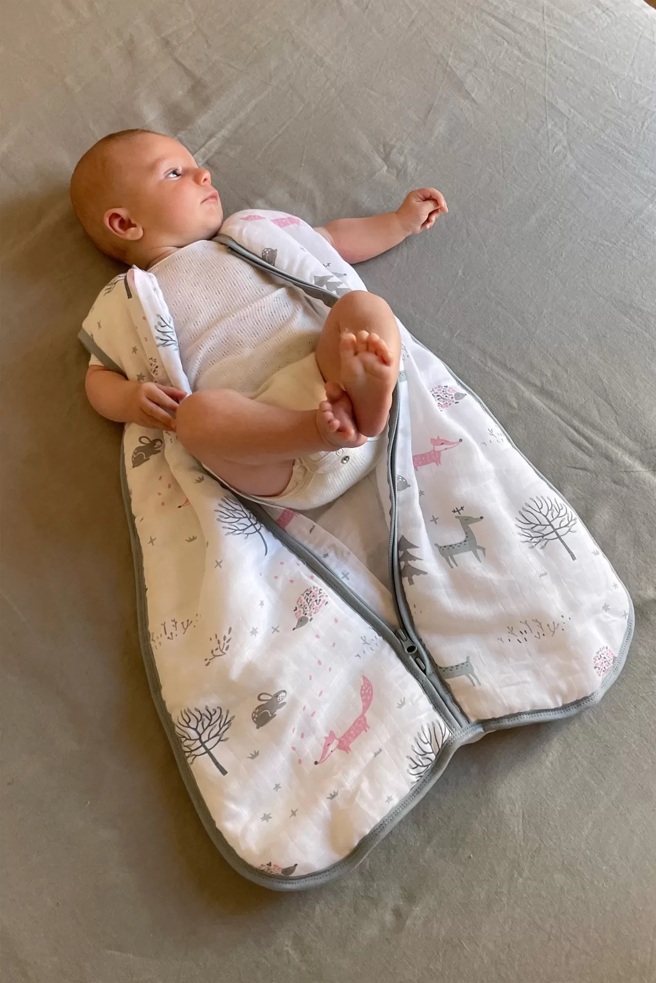 Saco de dormir para bebé branco com padrão de animais e árvores em cinza e rosa com fecho, com bebé deitado