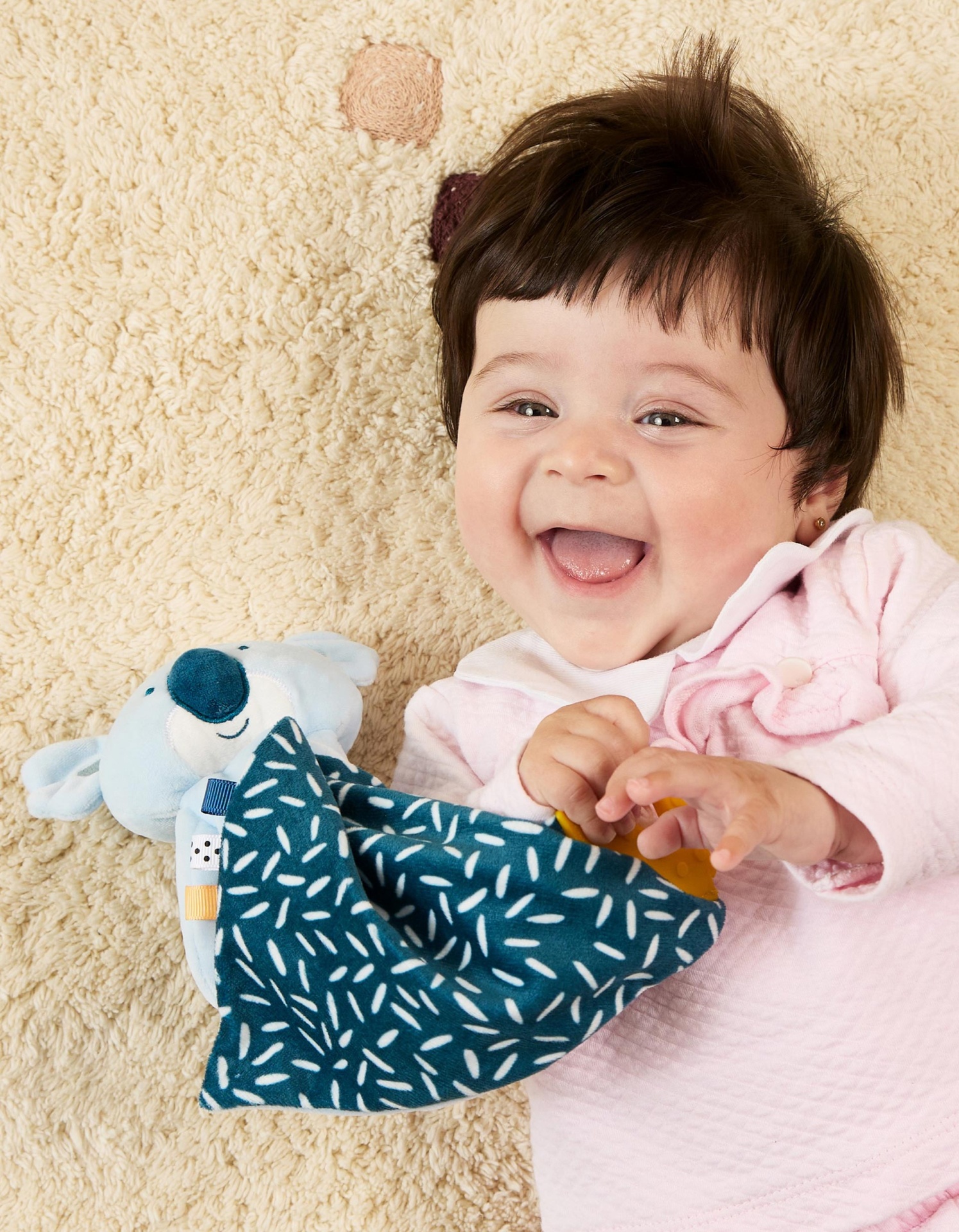 Bebé sorridente deitado com brinquedo de pelúcia azul e pano verde com padrão branco