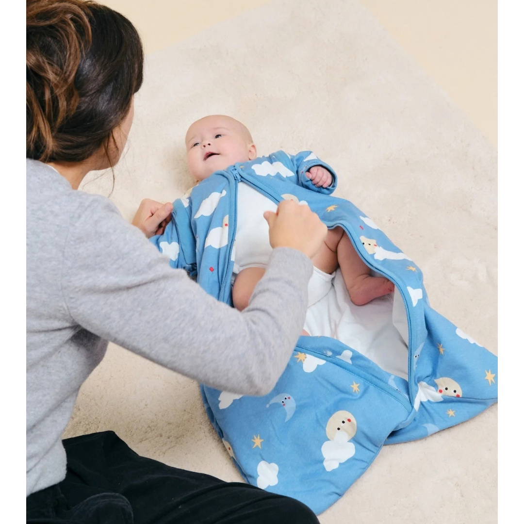 Bebé num saco de dormir azul com padrões celestes a ser ajustado por uma mulher com camisola cinza.