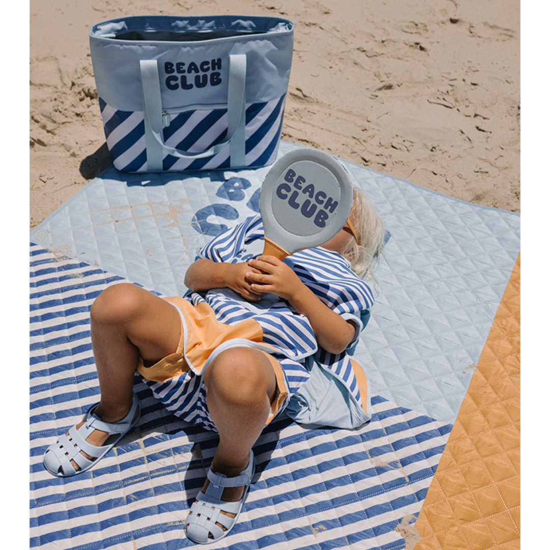 Criança sentada numa manta azul e branca segurando raquete BEACH CLUB junto a bolsa de praia