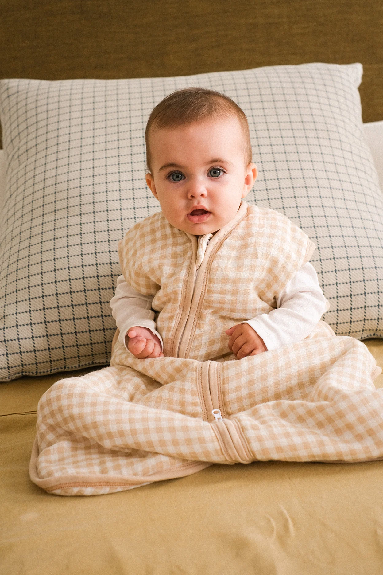 Bebé com saco de dormir bege e branco sentado em cama com lençol amarelo e almofada de padrão azul