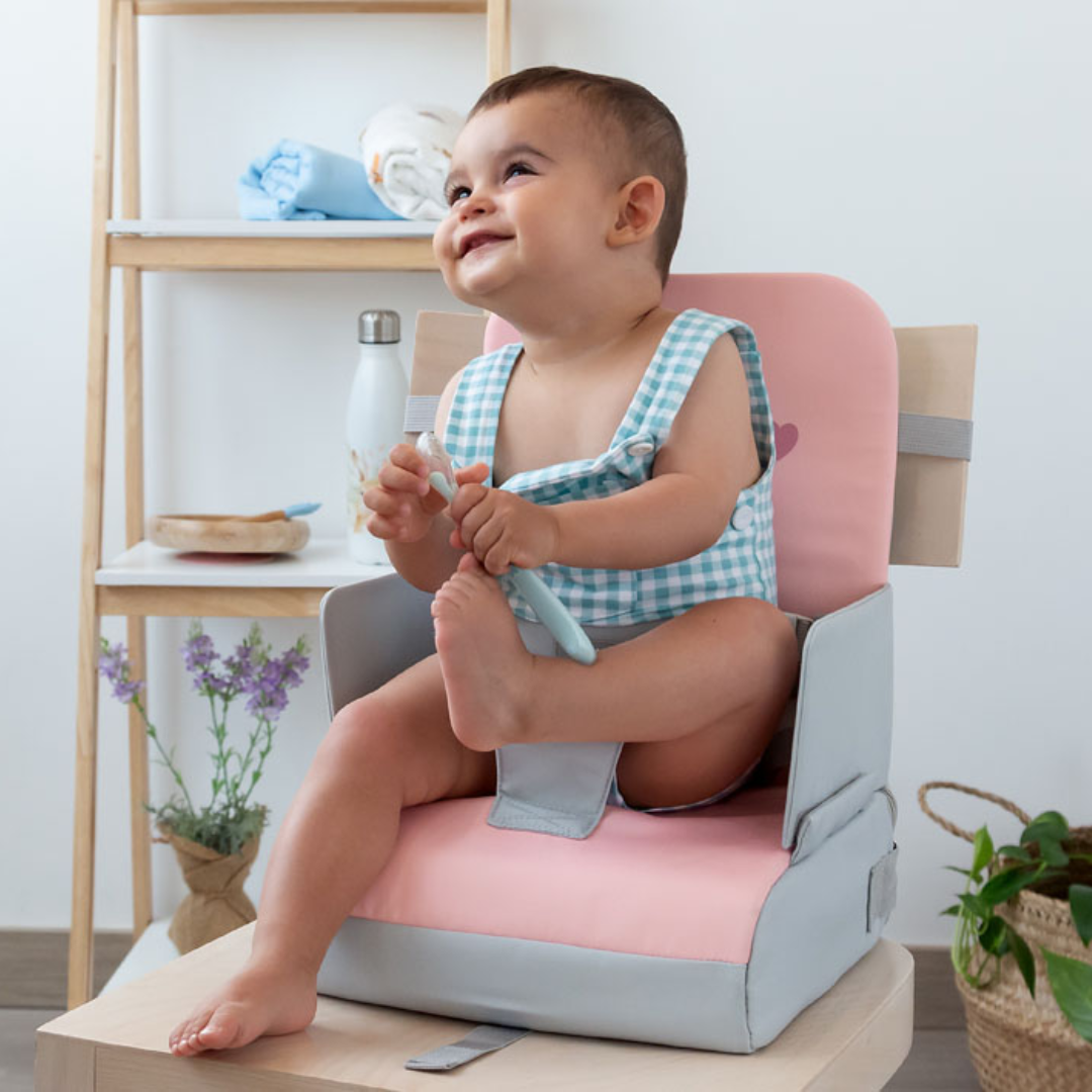 Bebé na cadeira almofadada cor-de-rosa e cinza, roupa xadrez azul e branca, segurando colher azul clara