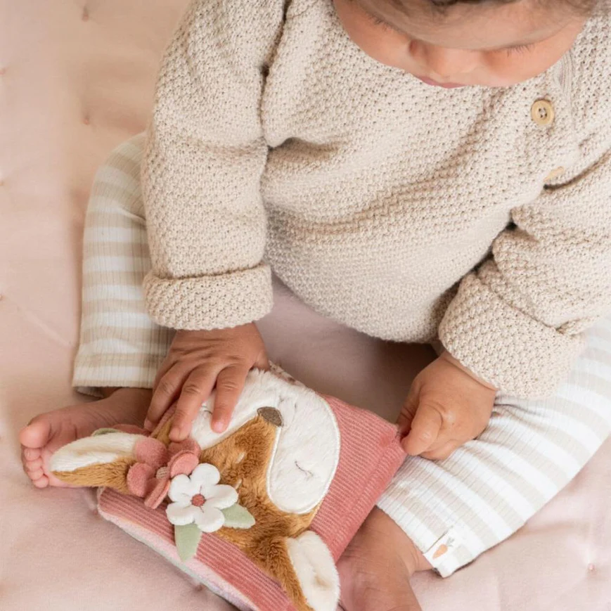 Criança com camisola de malha bege e calças listradas a tocar em livro de peluche cor-de-rosa com raposa e flores.