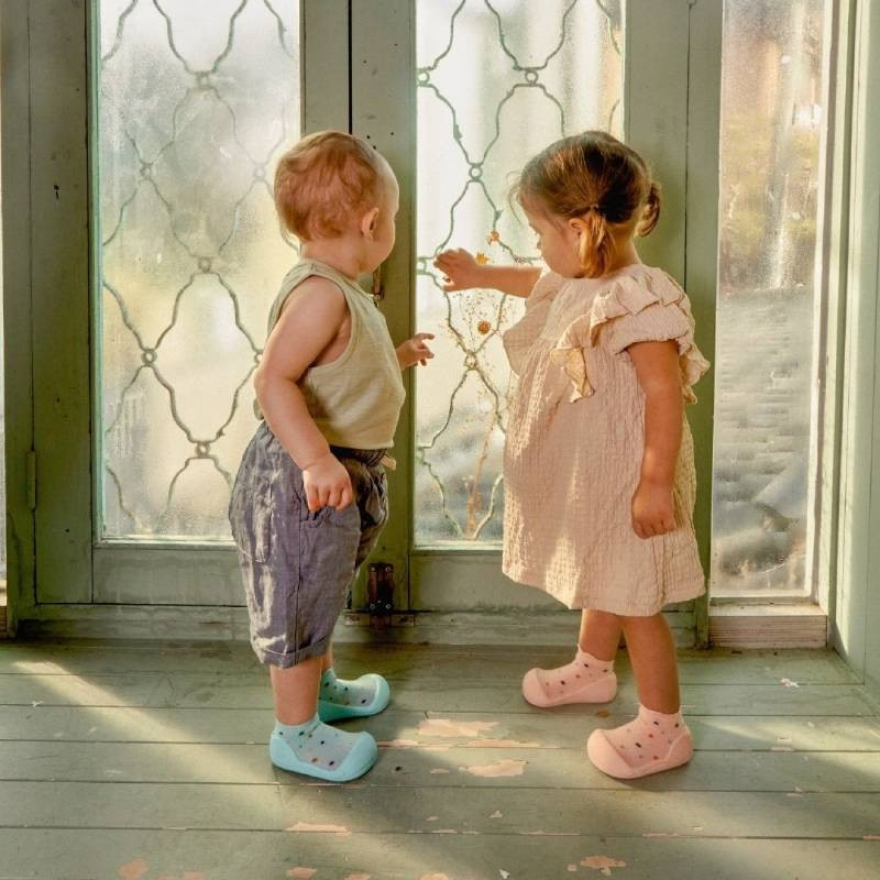 Duas crianças em pé numa varanda com janela texturizada, uma com calções cinza e sapatinhos azuis, outra com vestido rosa e sapatinhos rosa