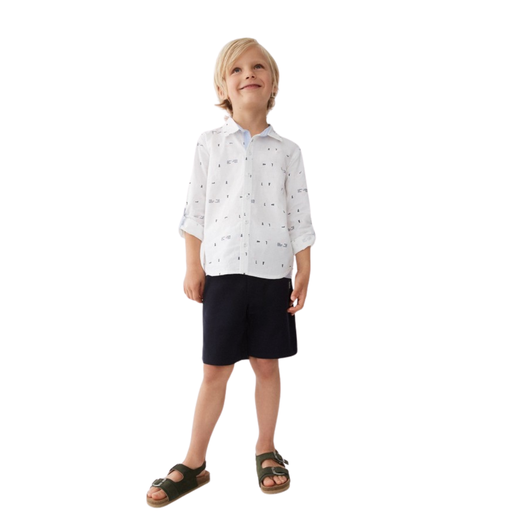 Criança com camisa branca de padrão de letras pretas, calções azuis escuros e sandálias verdes.