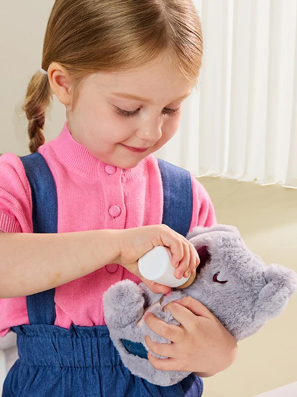 Criança com camisola rosa e vestido azul a alimentar peluche cinzento com uma mamadeira.