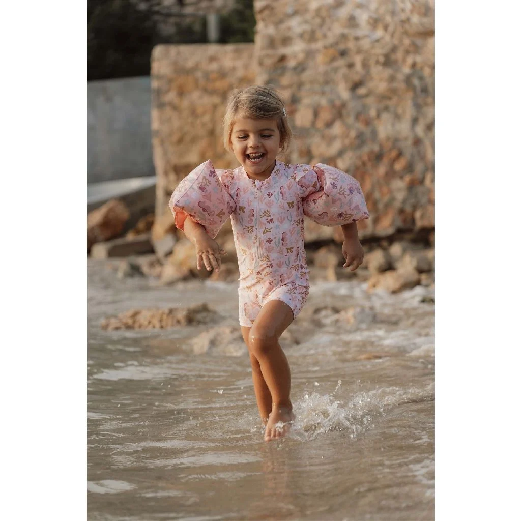 Criança com fato de banho rosa e manguitos a brincar na água numa praia.