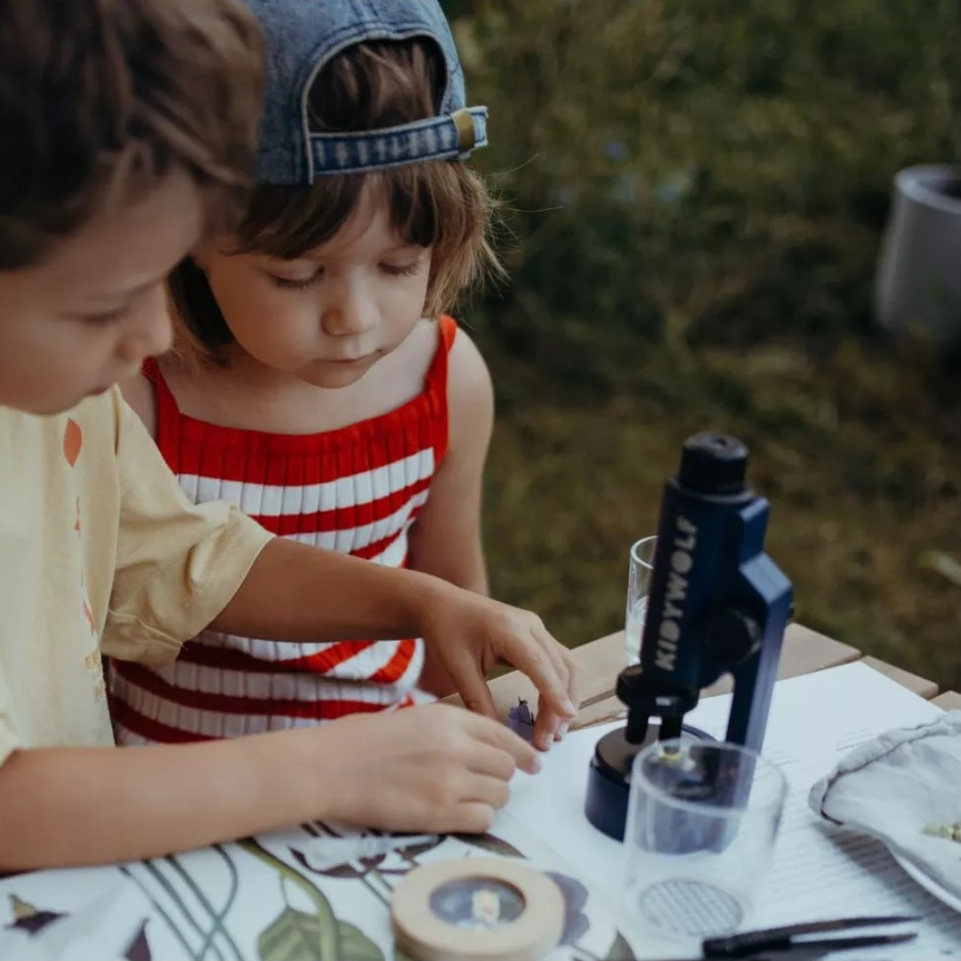 Crianças a usar microscópio azul escuro Dobex numa mesa com materiais de estudo ao ar livre