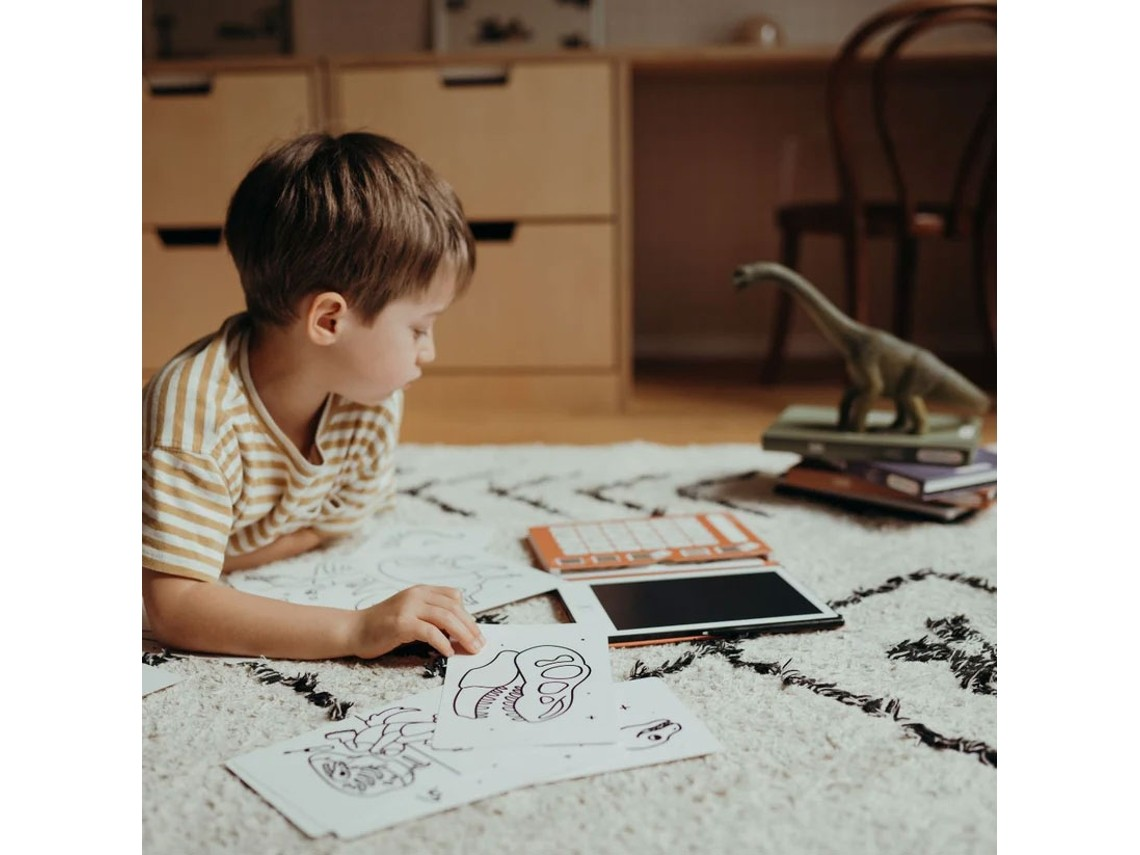 Menino deitado numa carpete a olhar para desenhos, com tablet e livros e um dinossauro de brinquedo ao lado