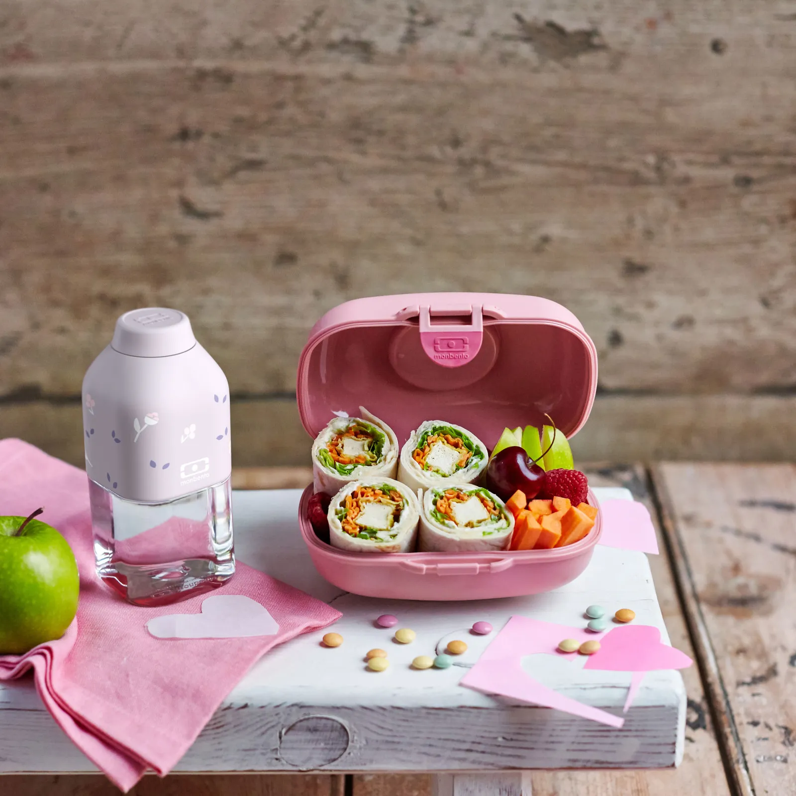 Marmita rosa com wraps, frutas e garrafa em mesa com fundo de parede texturada