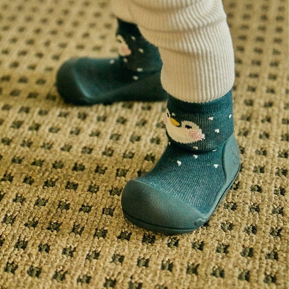 Meias azuis com desenho de pinguim e solado antiderrapante usadas por criança sobre tapete quadriculado.