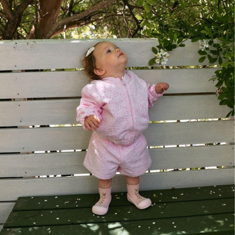 Criança com roupa rosa clara e sapatinhos bege junto a banco branco e flores